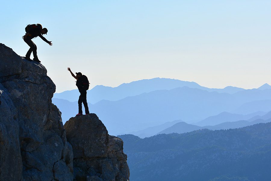 About Our Agency - Hikes Helping Each Other Climb a Rocky Mountain With More Mountains in the Background