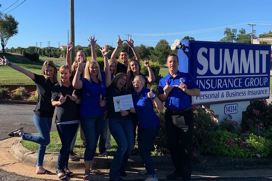 Community Involvement - Summit Insurance Group Inc Team Standing Together in Next to Their Company Signage Holding an Award and Cheering on a Sunny Day