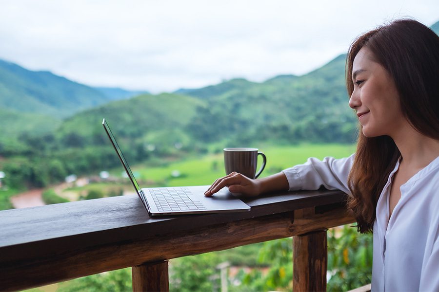 Blog - Woman Smiling and Staring at Her Open Laptop As She Sits Outside with Mountains in the Background